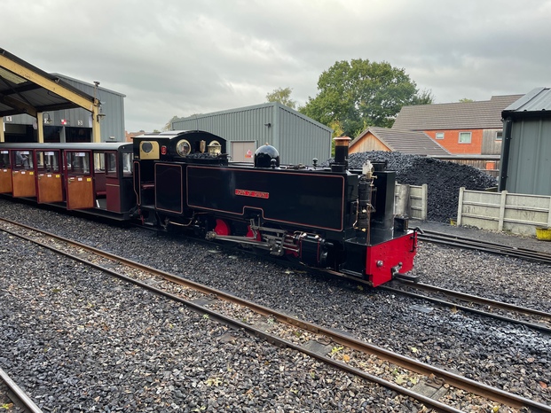 A black steam engine with a boxy shape, running on narrow rails. It has red fittings on the wheels and front bumper, and gold trim and decoration. It’s very shiny and well-cleaned! The cab is at the back of the train, a covered space with circular windows. It's pulling a single carriage, which is empty and has doors open towards the camera.